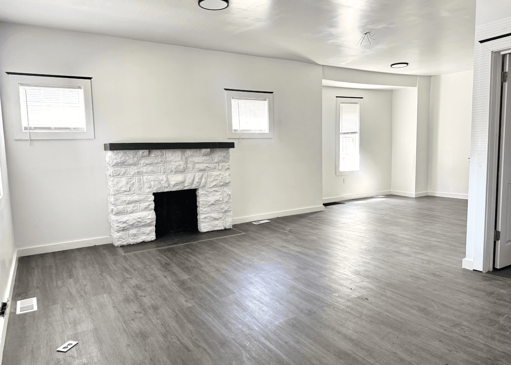Bright, empty living room with white walls, a white stone fireplace, several small windows, and gray wood-look flooring.