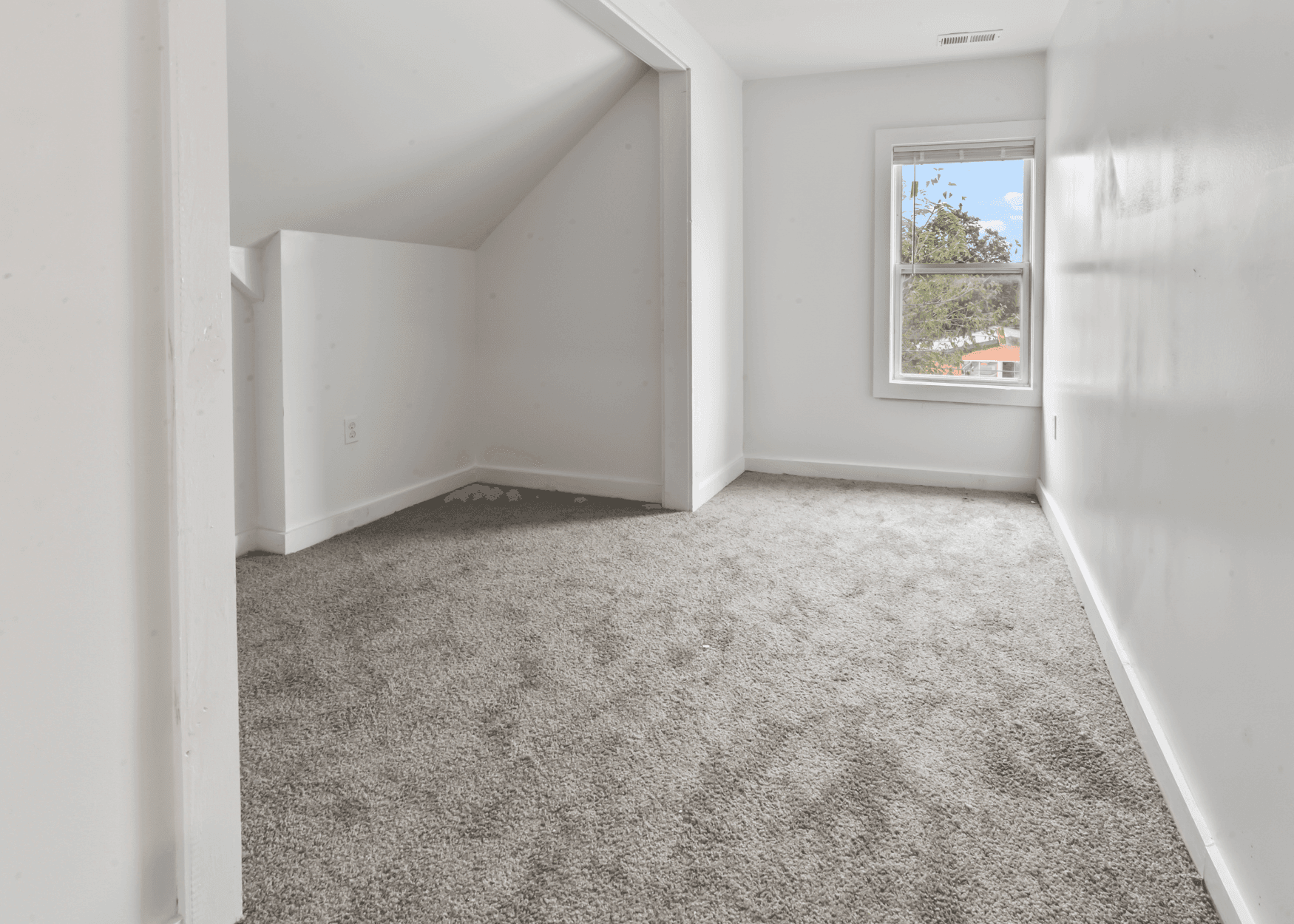 Empty white room with a beige carpet, angled ceiling nook, and a window showing trees outside.