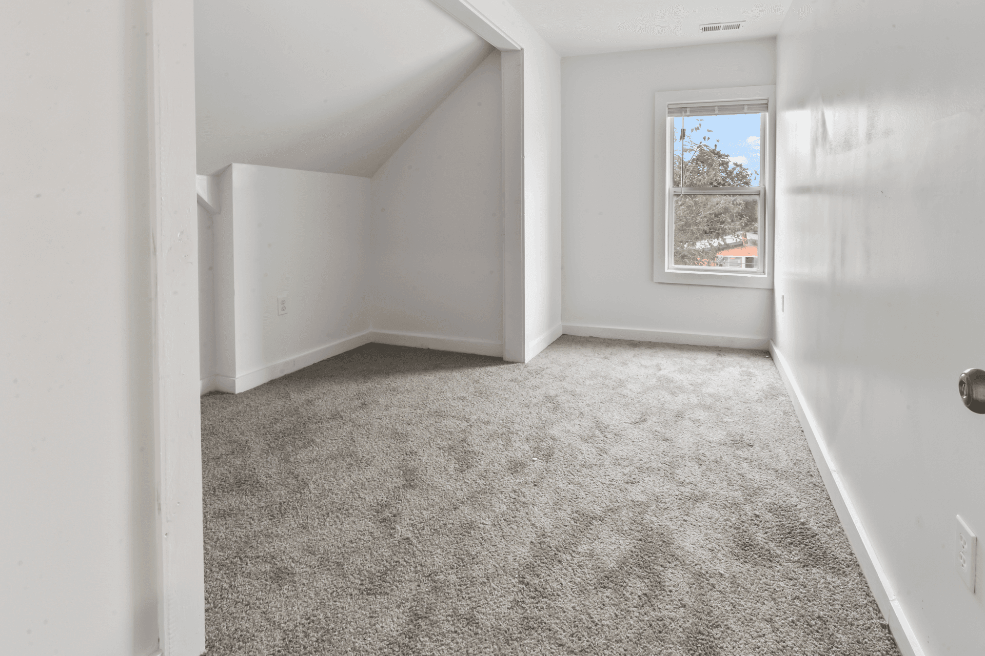 Small bright attic room with a sloped ceiling, white walls, and gray carpet, featuring a window with a view of trees.