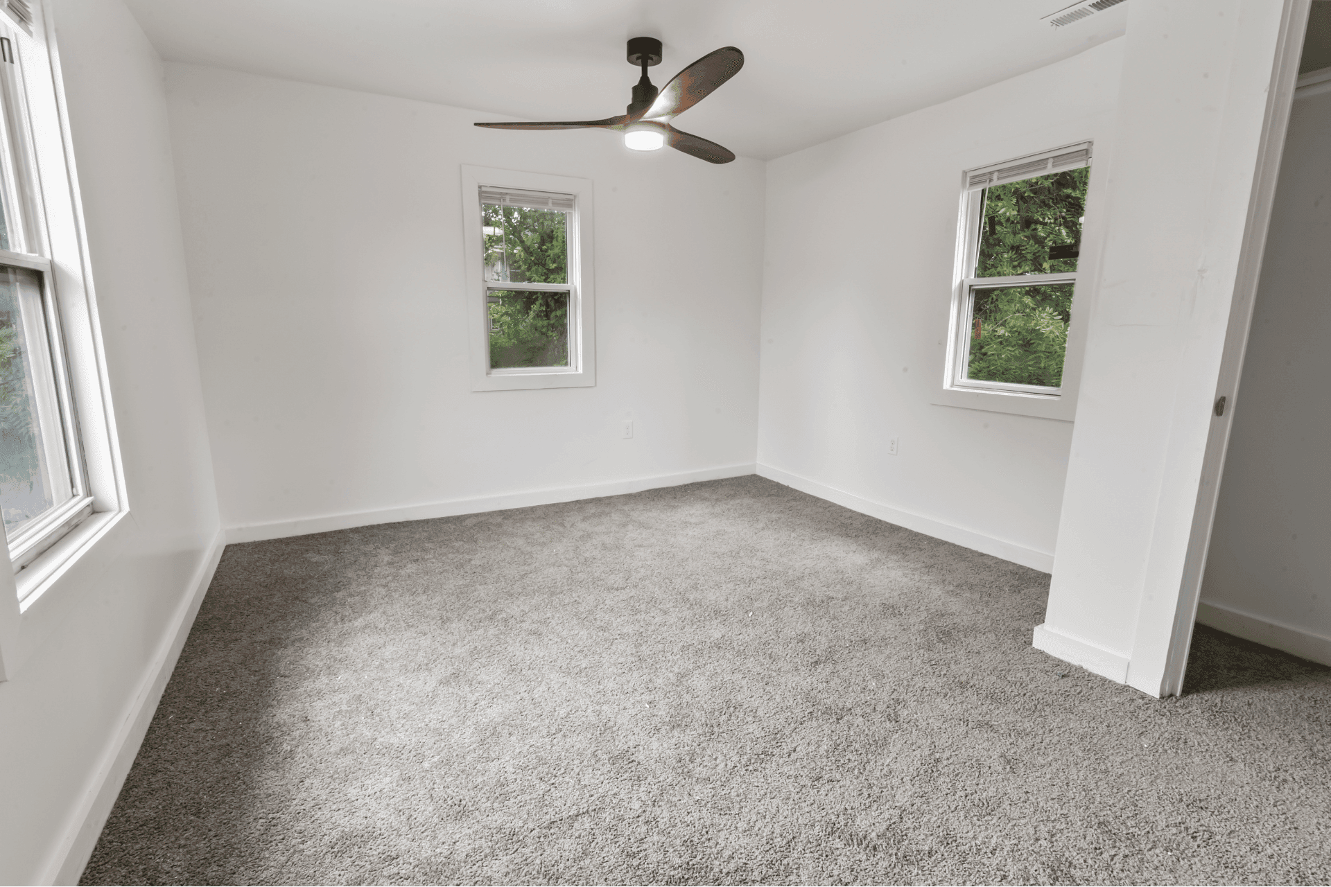 Empty white room with two windows, gray carpet, and a ceiling fan; view shows green trees outside