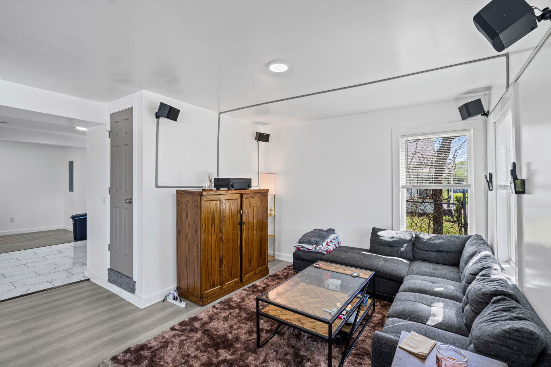 Living room with a gray sectional sofa, brown rug, glass coffee table, and wall-mounted speakers around a windows view.