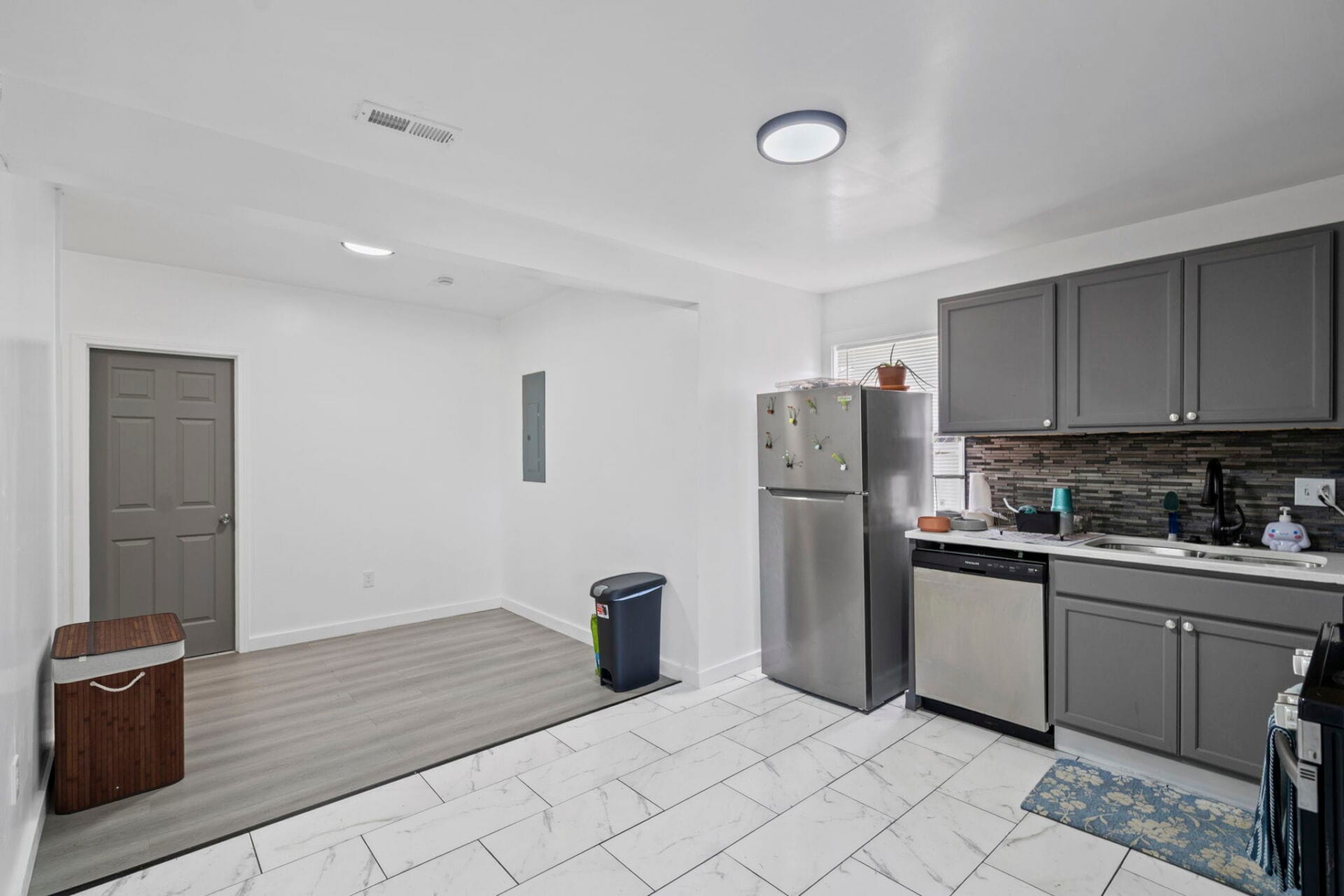 Modern kitchen with gray cabinets, stainless steel refrigerator and dishwasher, and white marble-look floor tiles.