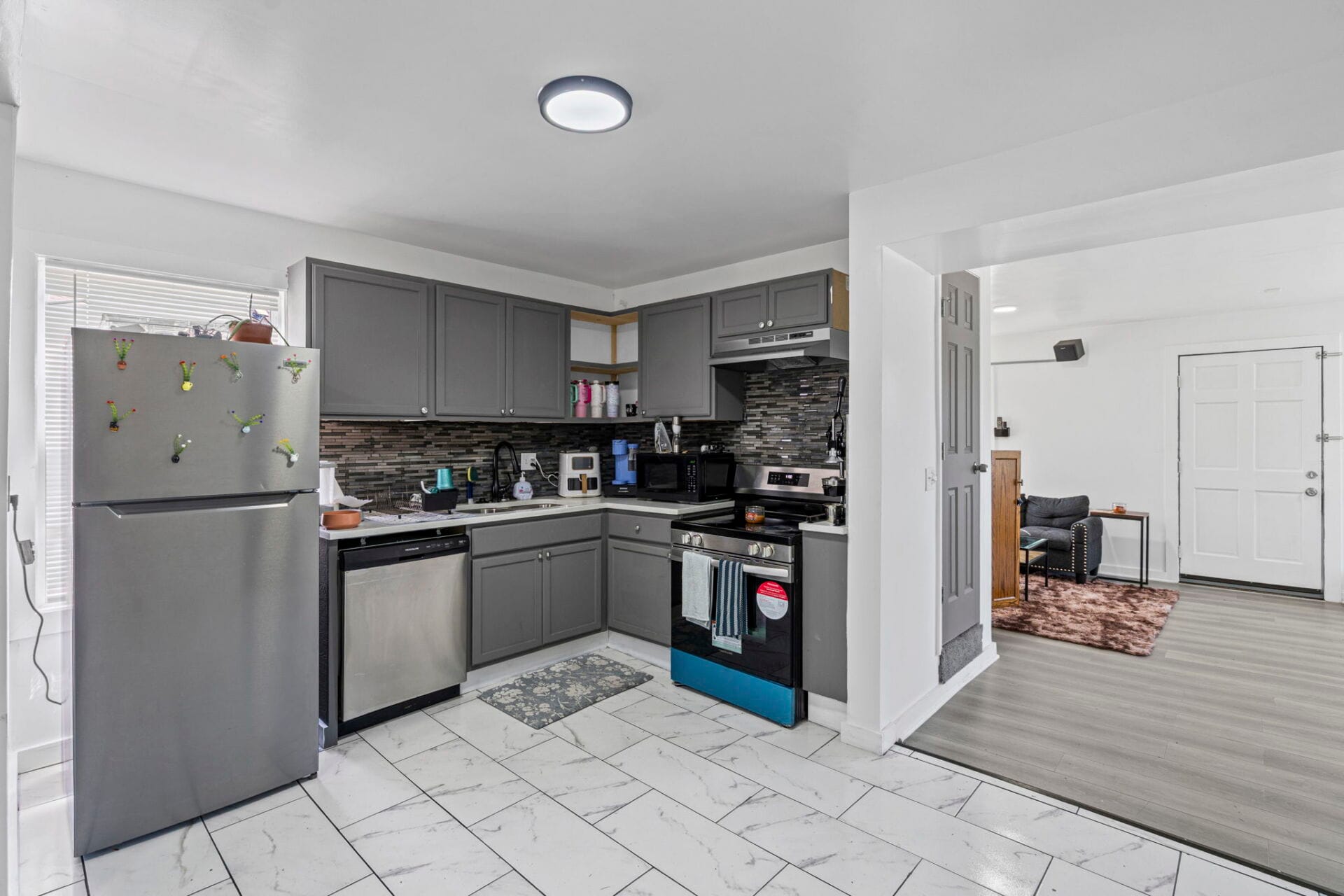Modern grey kitchen with stainless steel fridge, black backsplash, and stainless appliances beside an open living area doorframe to the right.