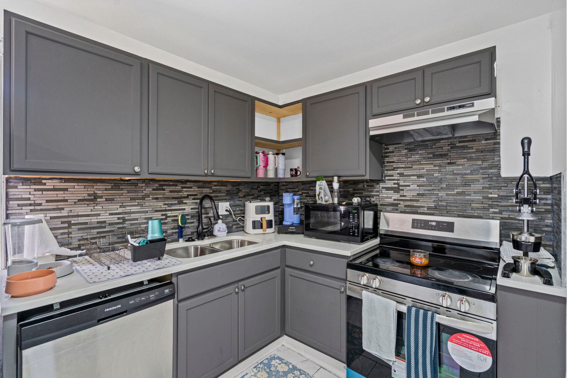 L-shaped kitchen with dark gray cabinets, stainless steel appliances (dishwasher, microwave, range) and a black-and-gray tiled backsplash under bright lighting; a double sink is visible.