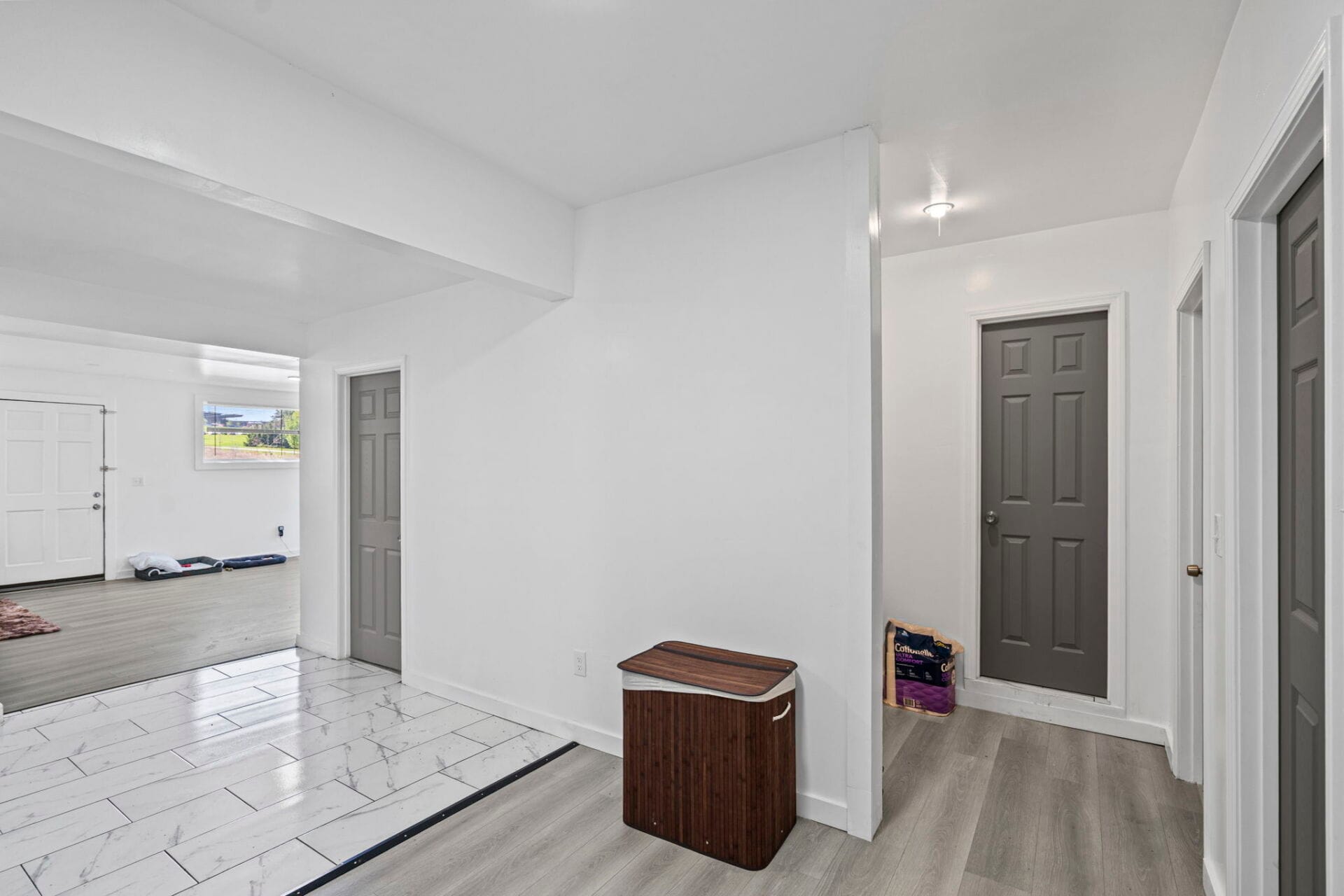 Bright interior hallway with white walls, gray doors, and a small window view outside; a wooden bench sits near the wall.