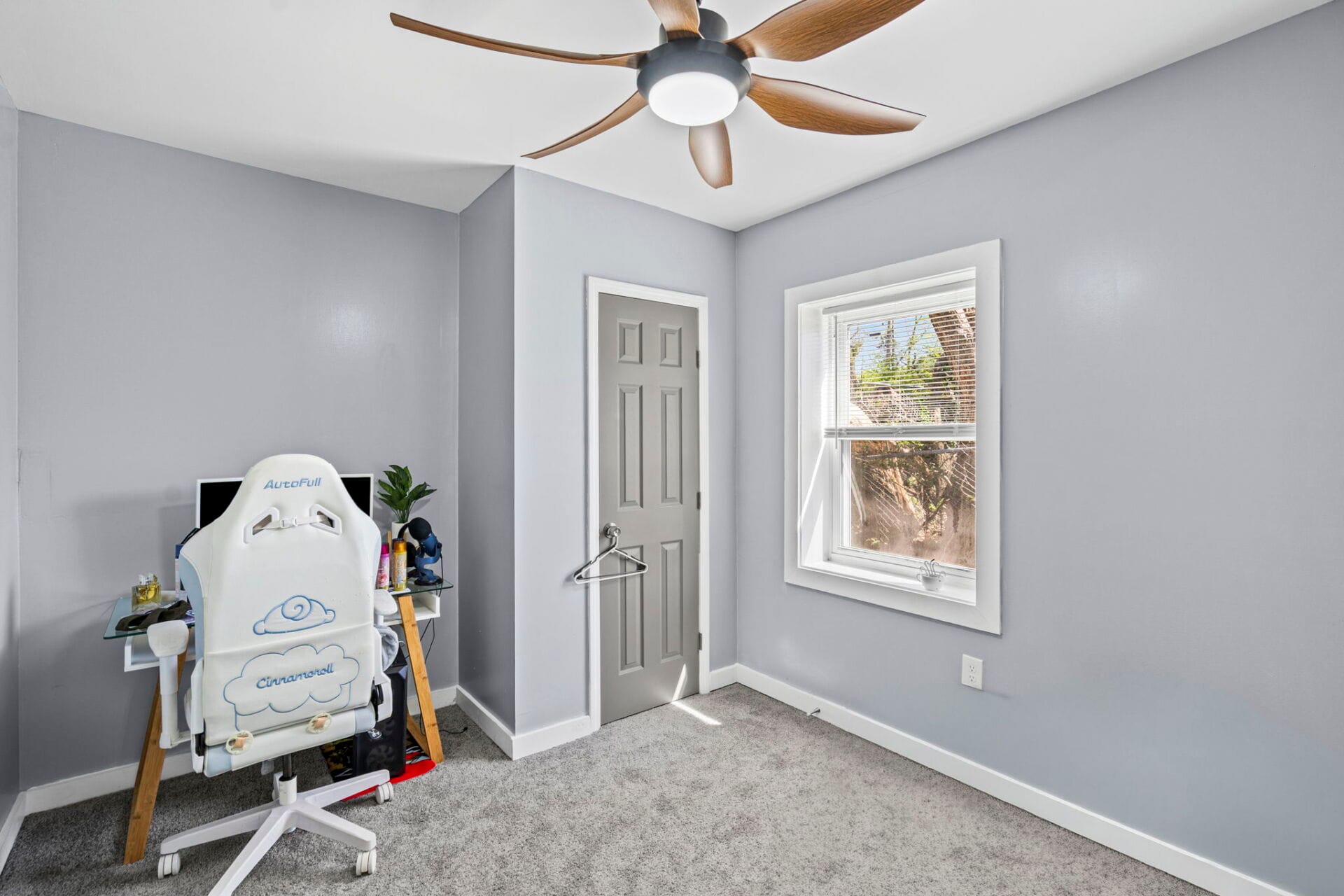 Home workspace with a white ergonomic chair at a glass desk, gray walls, and a window letting in sunlight.