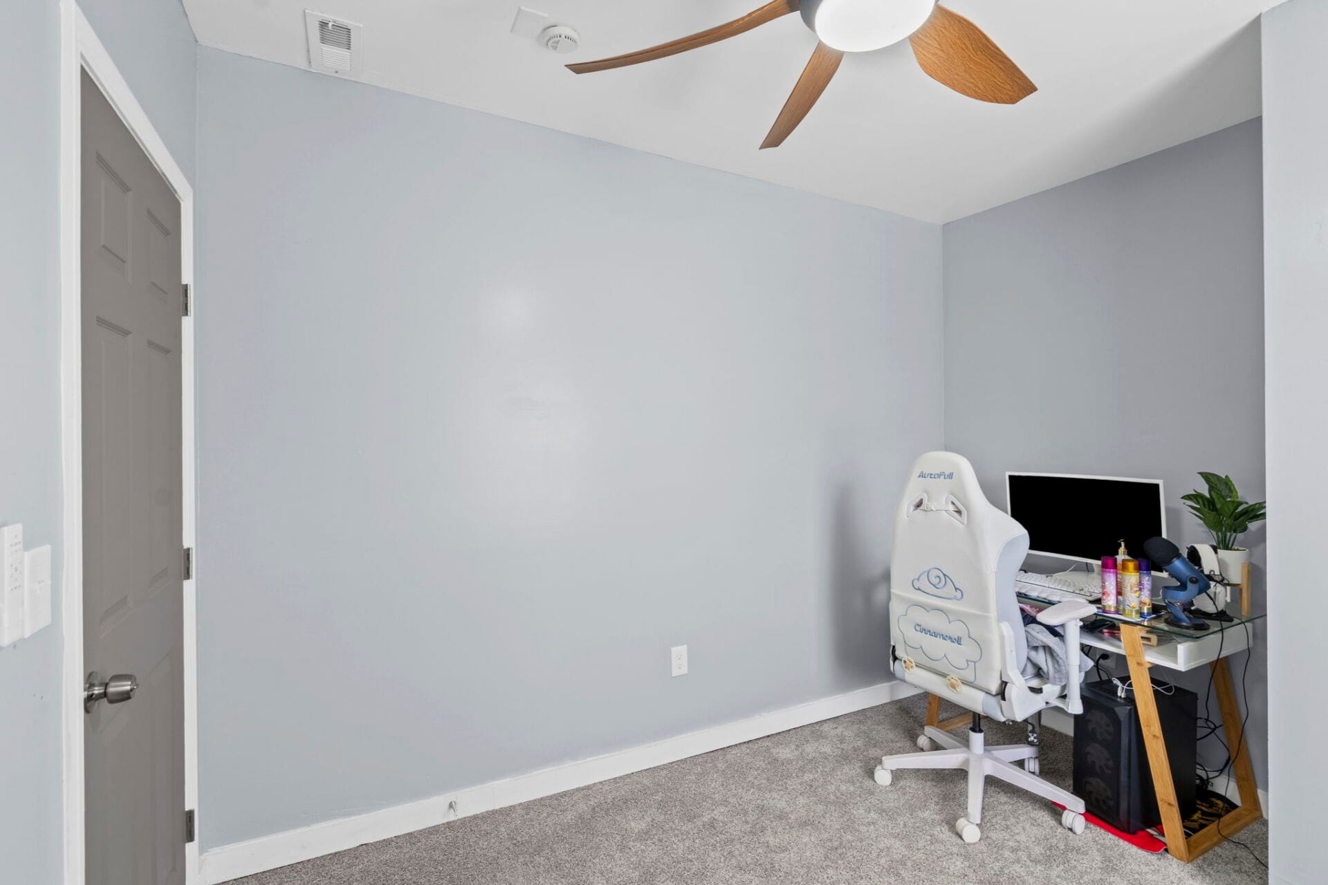 Home office corner with a light blue wall, gray door, and a white desk setup including a monitor and gaming chair.