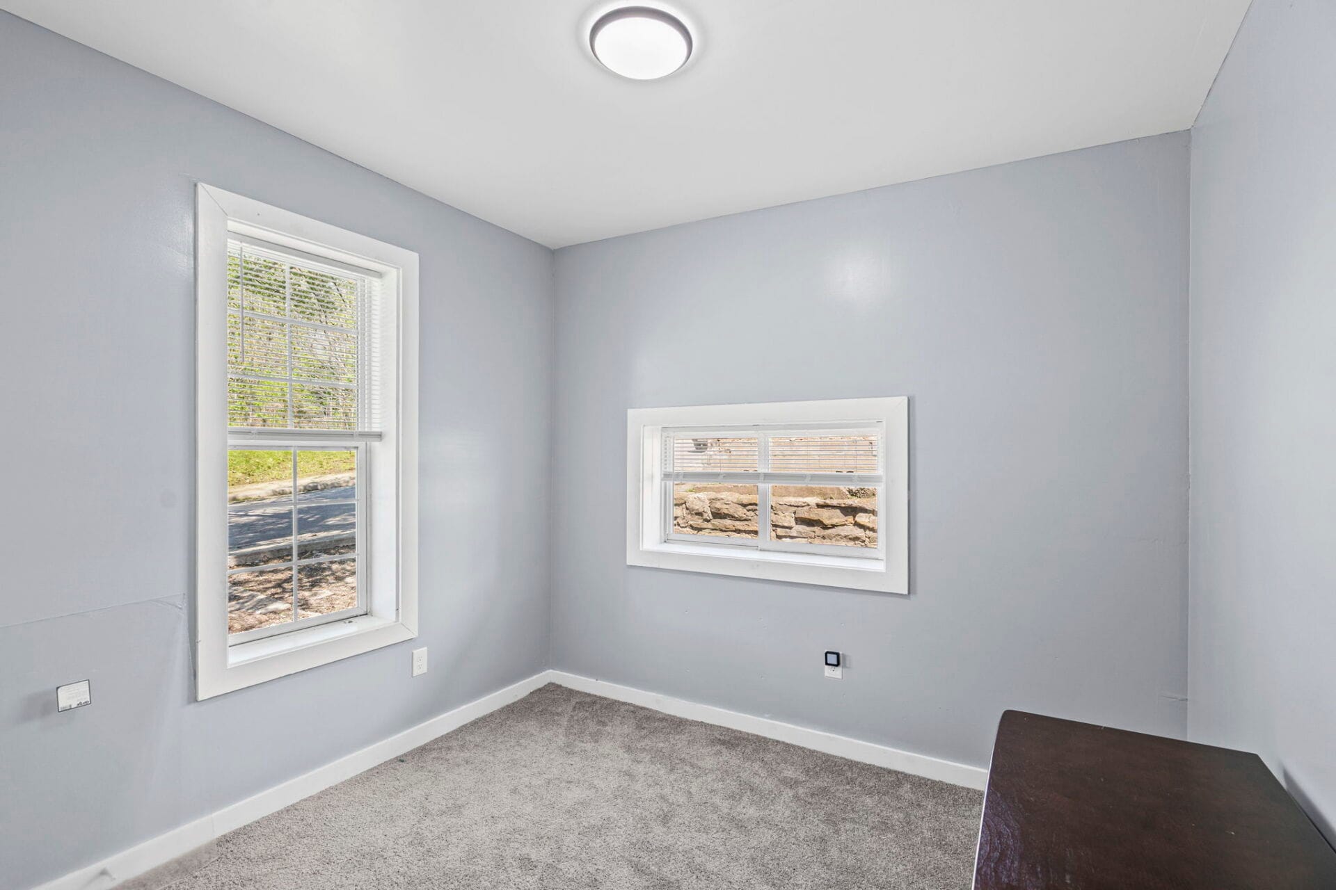 Empty room with light gray walls, two white-framed windows (tall left and small right), and beige carpet.