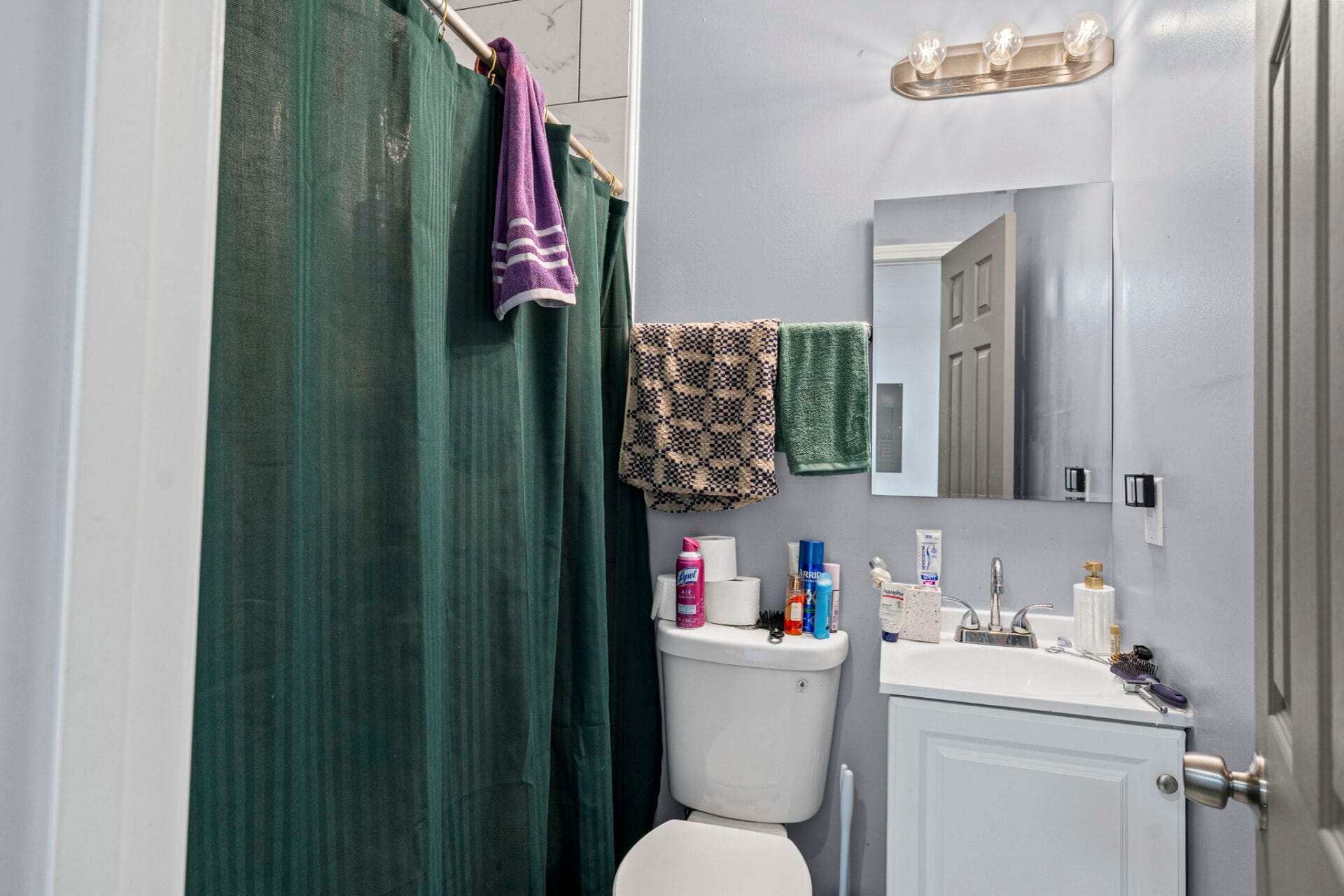 Small bathroom with a dark green shower curtain, purple towel, beige checkered towel, and a green towel on a rack; mirror above sink and counter cluttered with toiletries.