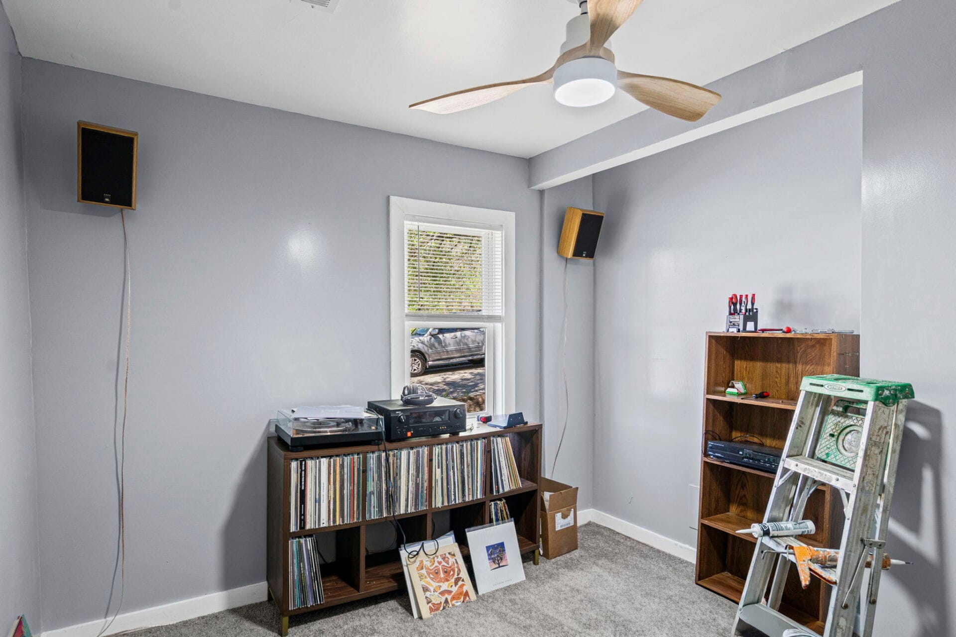 Corner of a small music room with gray walls, window, speakers on the walls, and a vinyl-record shelf filling the cabinet beneath a turntable setup.