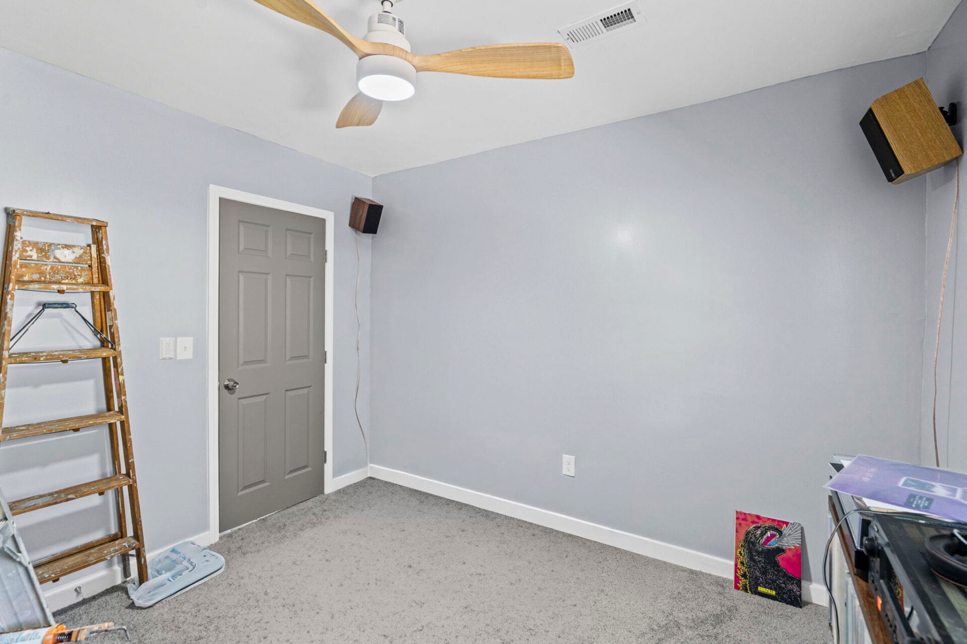 A small room under renovation with a wooden ladder on the left, gray walls, and a ceiling fan; speaker mounted on the right wall and a door in the corner.