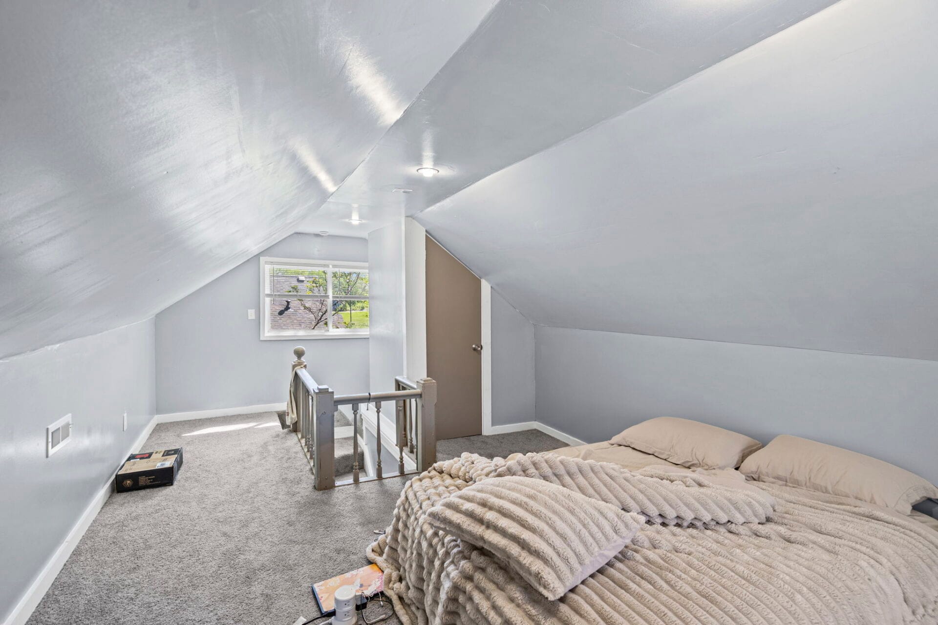 Attic bedroom with sloped ceiling, gray walls, and a bed on the right; a small window and staircase railing at the far end.