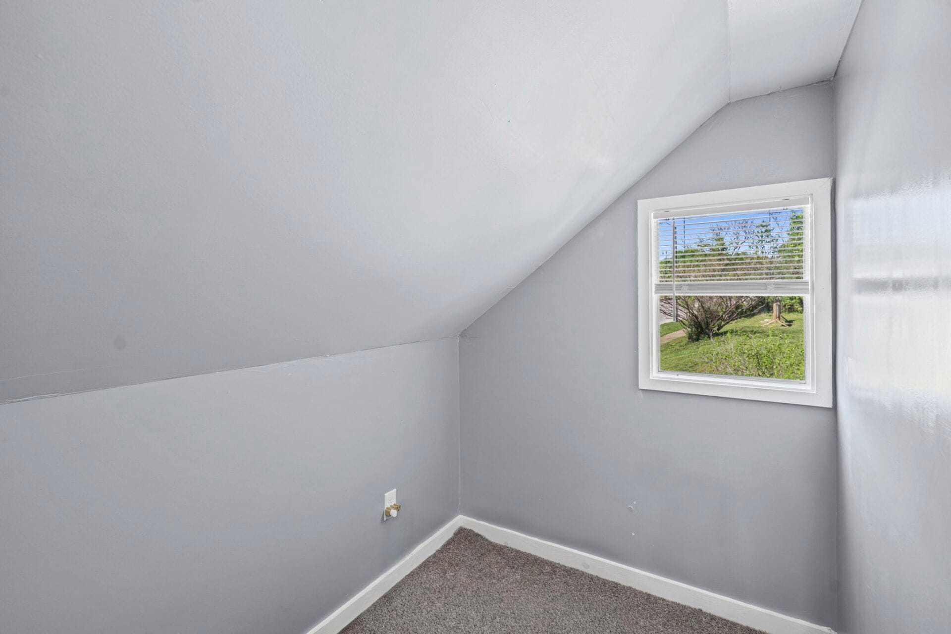 Small gray room with a sloped ceiling and a white-framed window with blinds; outside shows trees and grass.