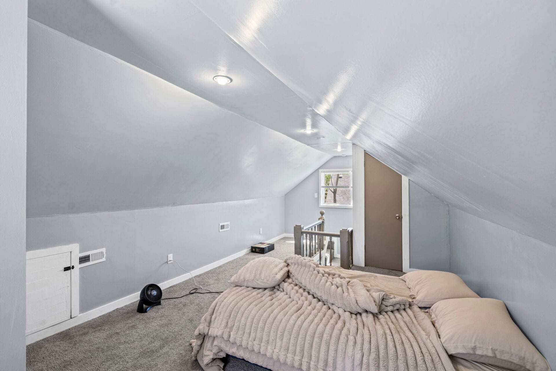 Cozy attic bedroom with sloped ceilings, a bed with beige comforter in the foreground, and a small window at the far wall.