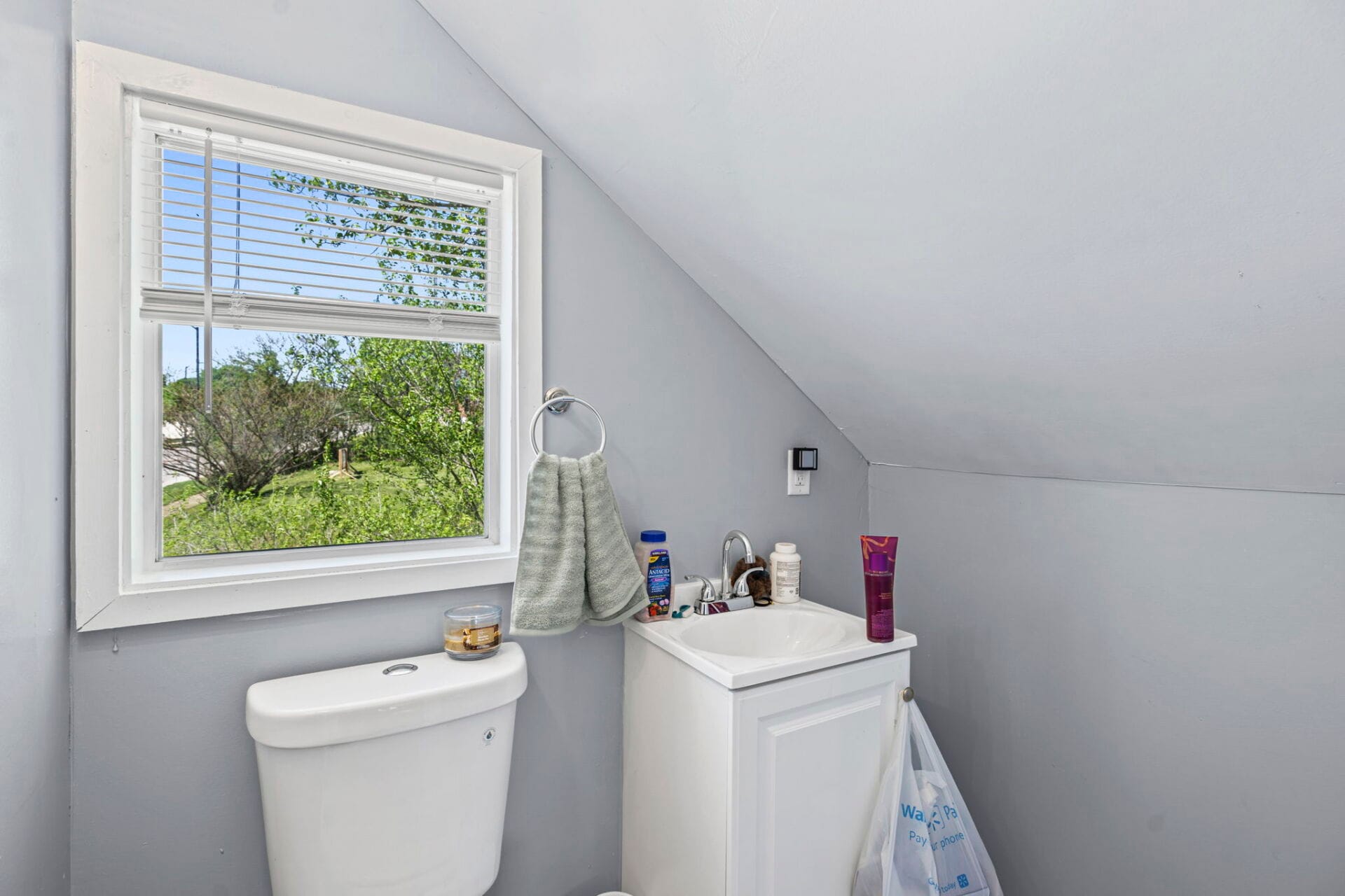 Small bathroom with a window, towel ring, sink, and a stacked washer beneath a sloped ceiling; toiletries on the counter appear in view.