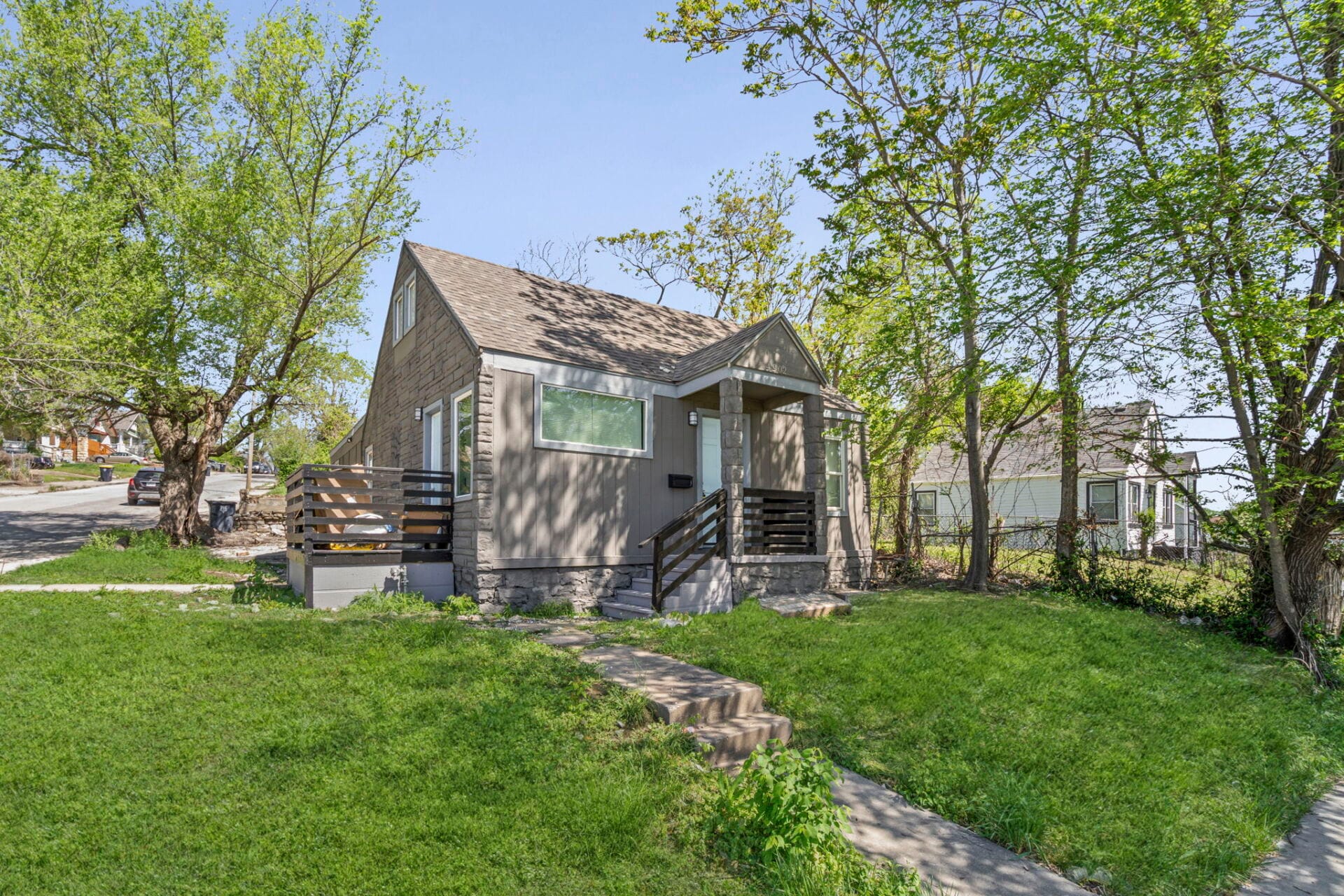 Small gray stone bungalow with a front porch, steps, and wooden railing, set in a green yard with trees.