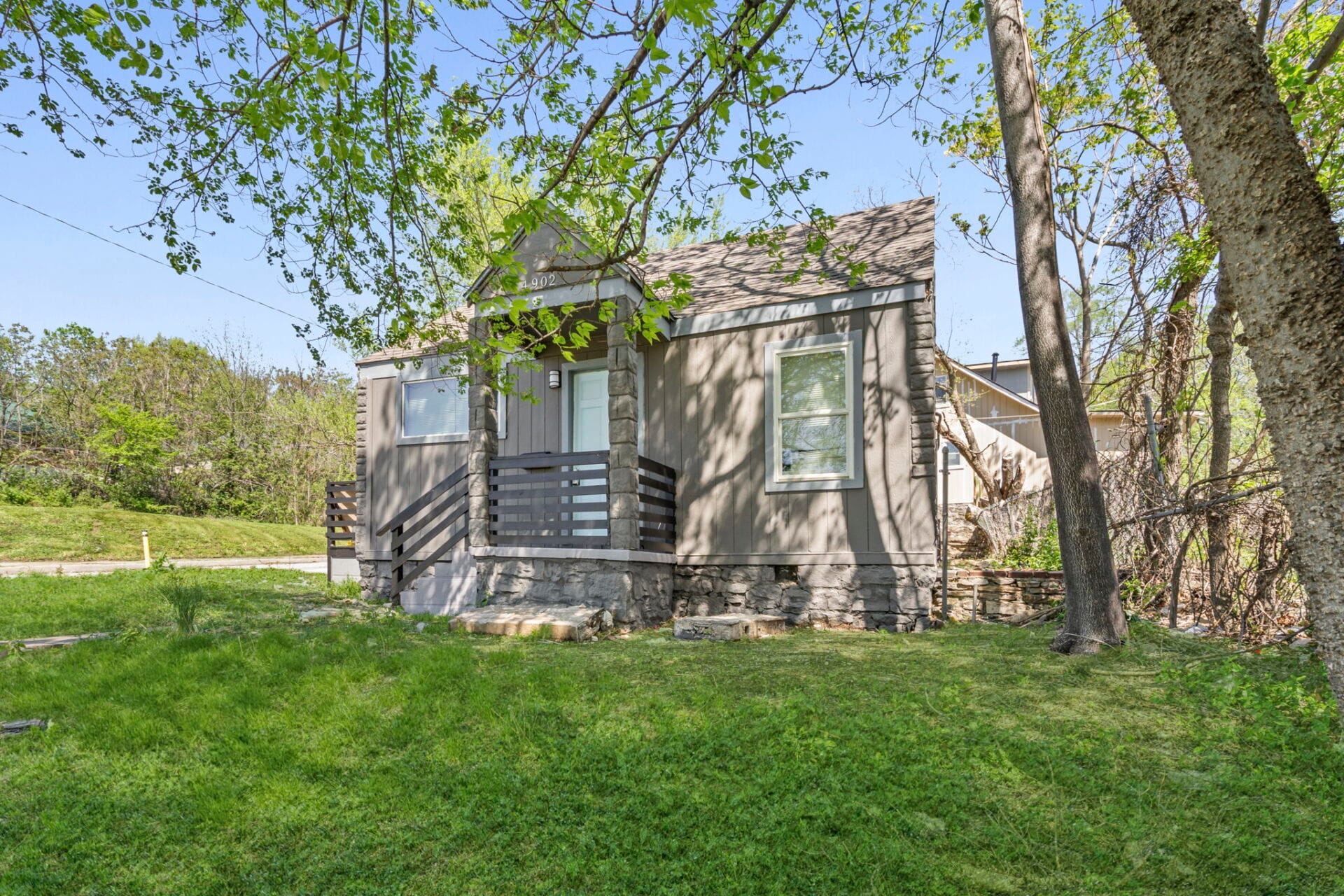 Small gray house with a stone foundation and front porch, surrounded by green grass and leafy trees on a sunny day.