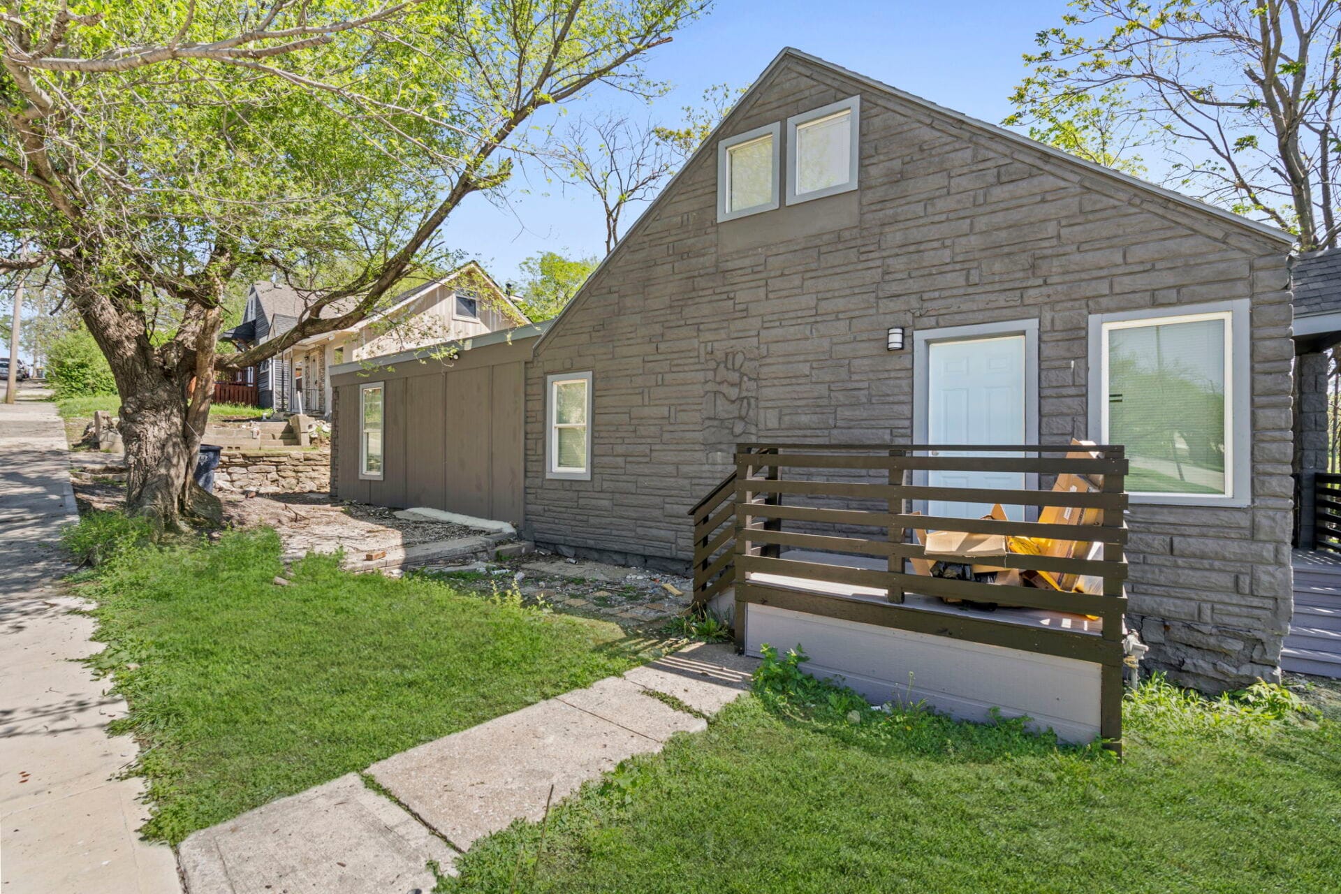 Stone-gray house with a steep, triangular roof and small front door, surrounded by a wooden railing and large tree branches overhead.