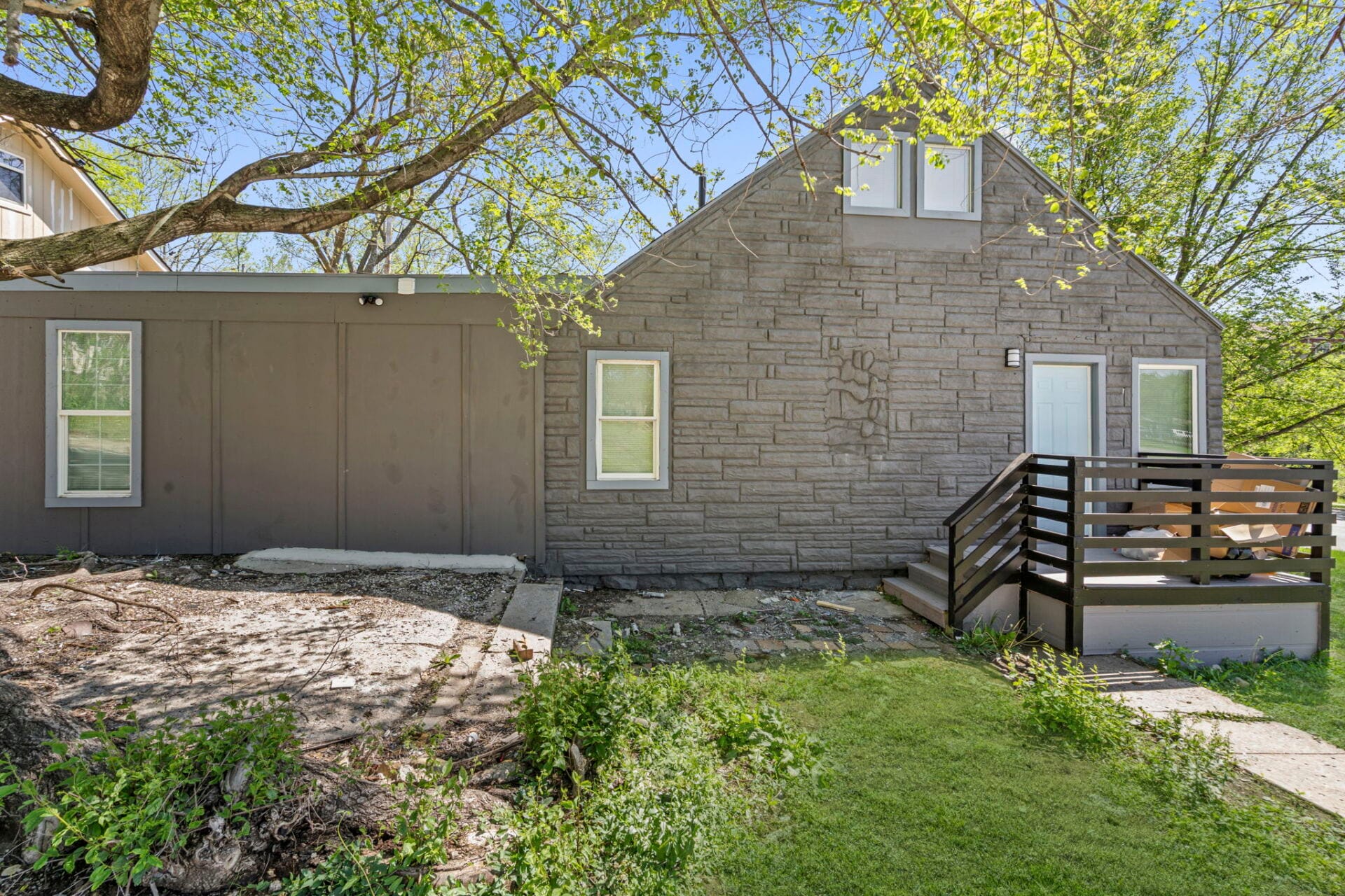 Gray stone exterior house with a small second-story dormer, blue door, and wooden deck on the right, under leafy tree branches.