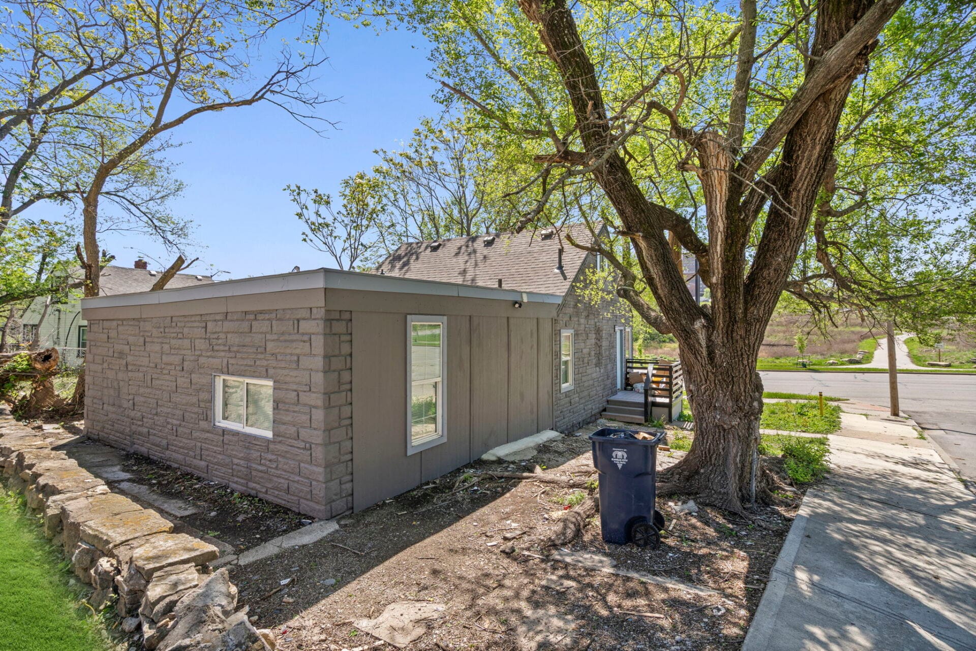Single-story gray brick house with a large tree in the front yard on a sunny day; curbside blue recycling bin nearby.