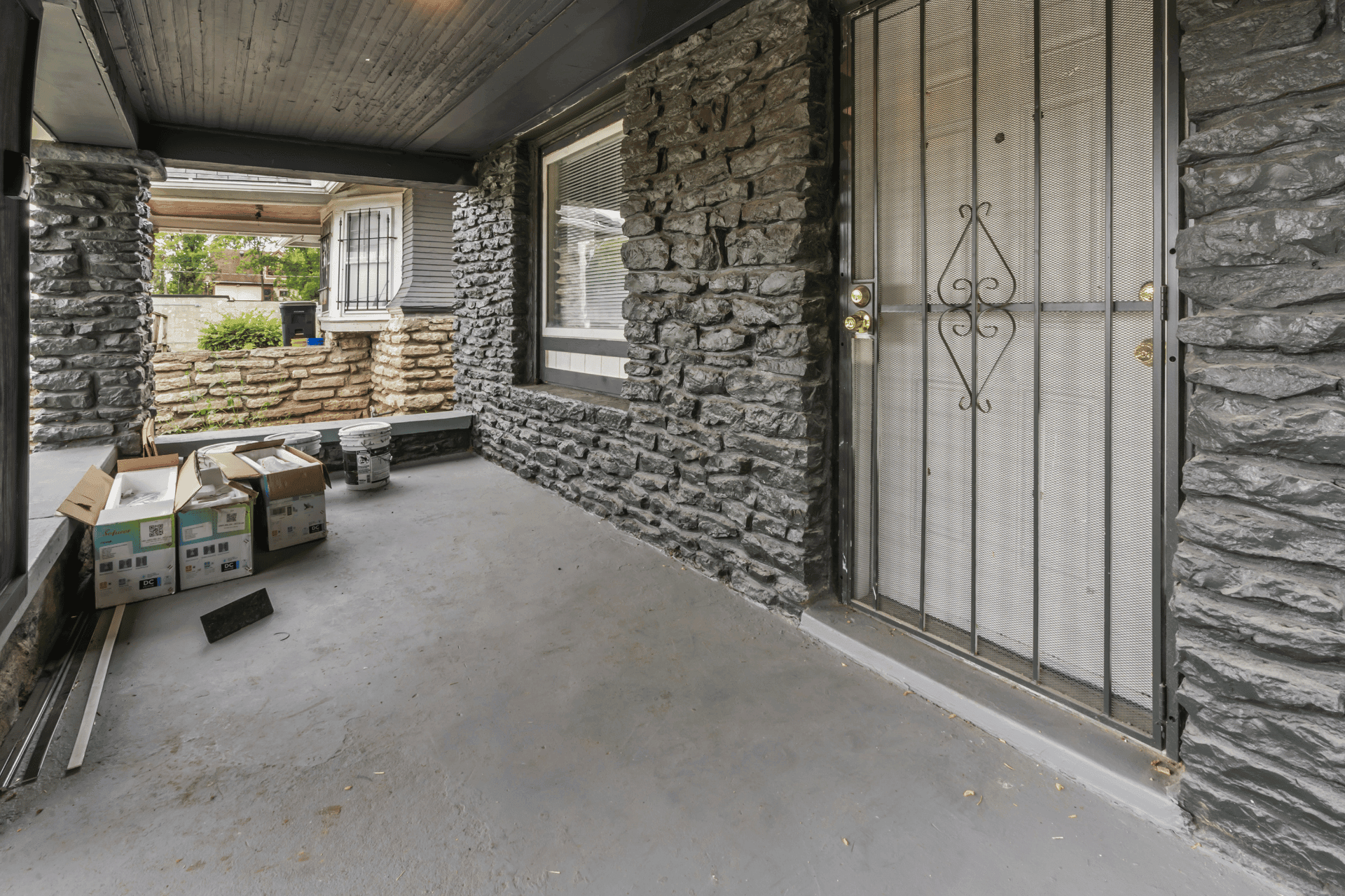 Covered stone-entry porch with a metal security door; boxes, buckets, and construction debris on the concrete floor.