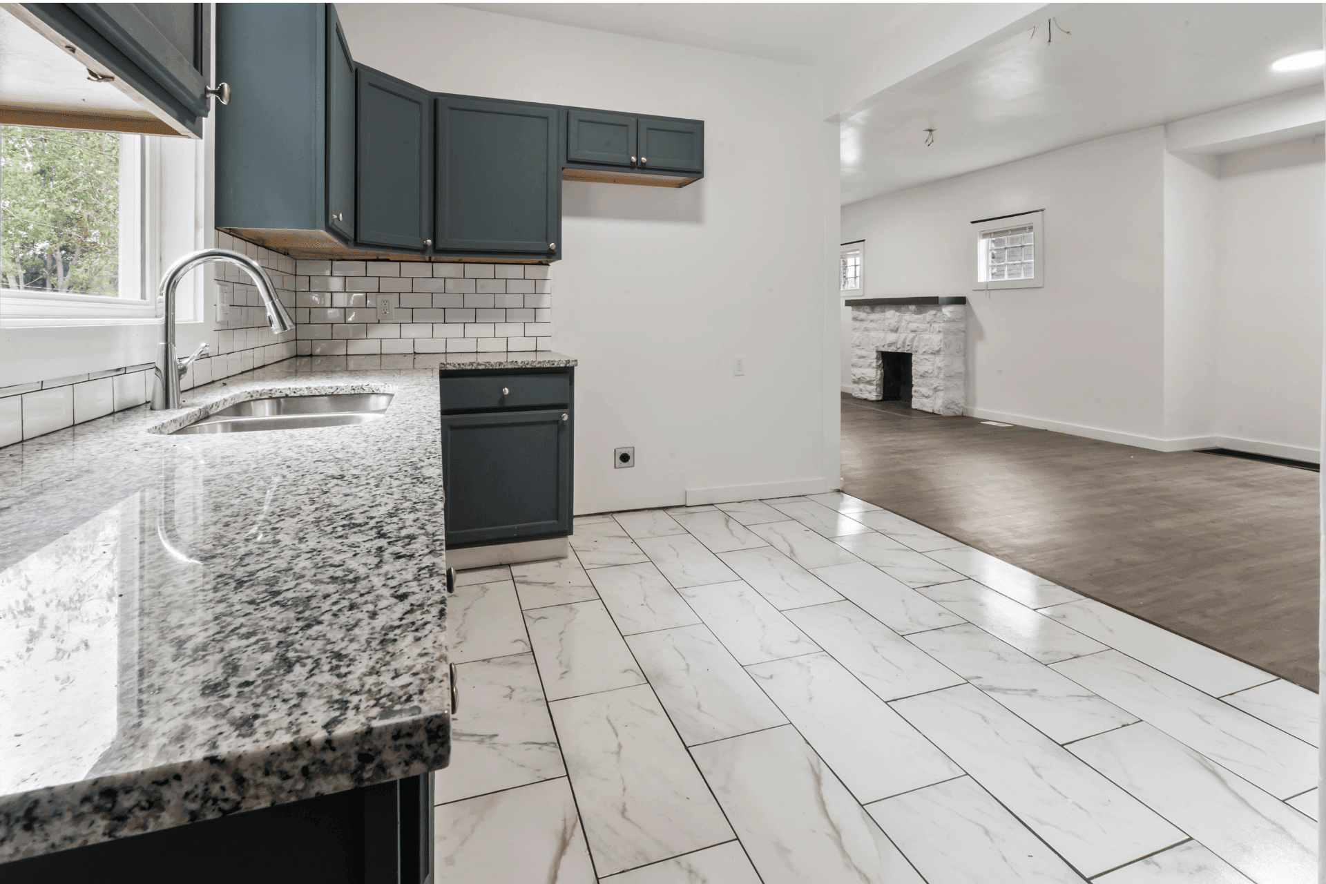 Modern kitchen with speckled granite countertops, dark blue cabinets, white subway tile backsplash, and a double sink; open to a living area with a stone fireplace.