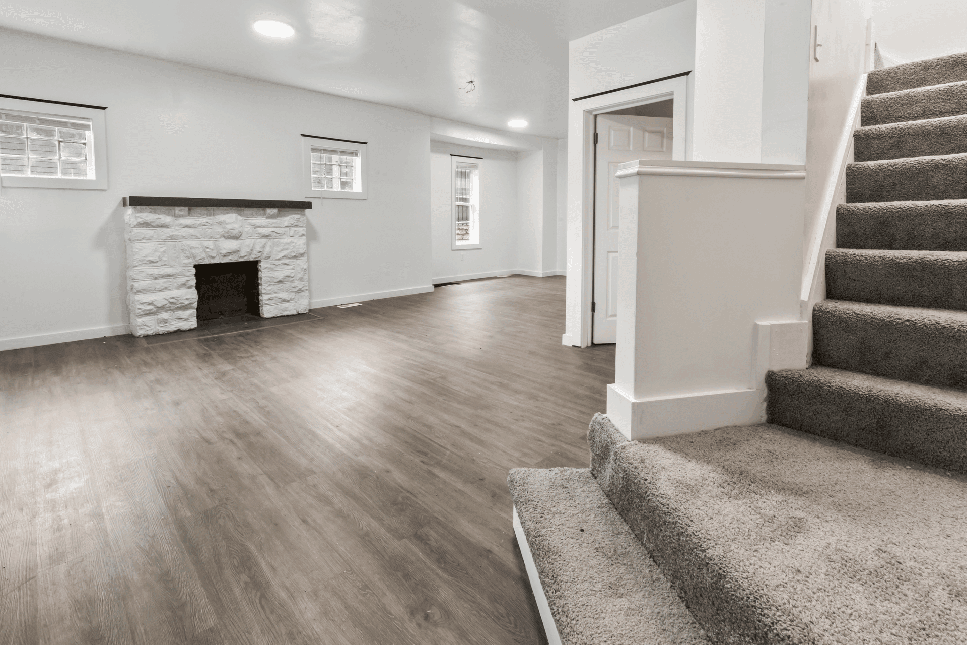 Finished basement living area featuring a white stone fireplace along the left wall, wood-look flooring, and multiple small windows; carpeted staircase on the right.