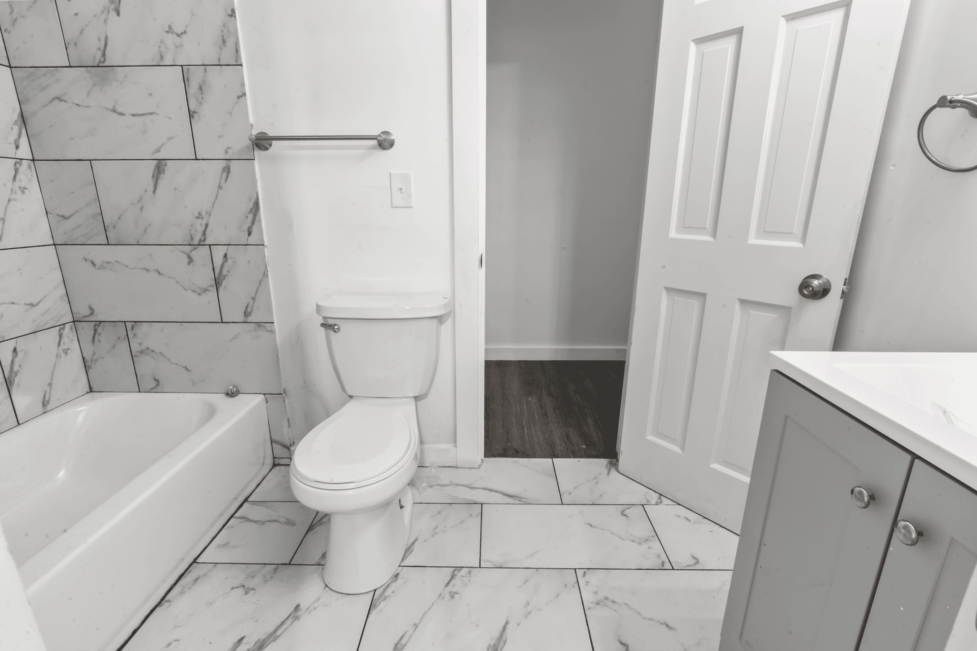 White bathroom with marble tile walls and floor, toilet beside a bathtub, towel bar on the wall, and an open door revealing a hallway to the right