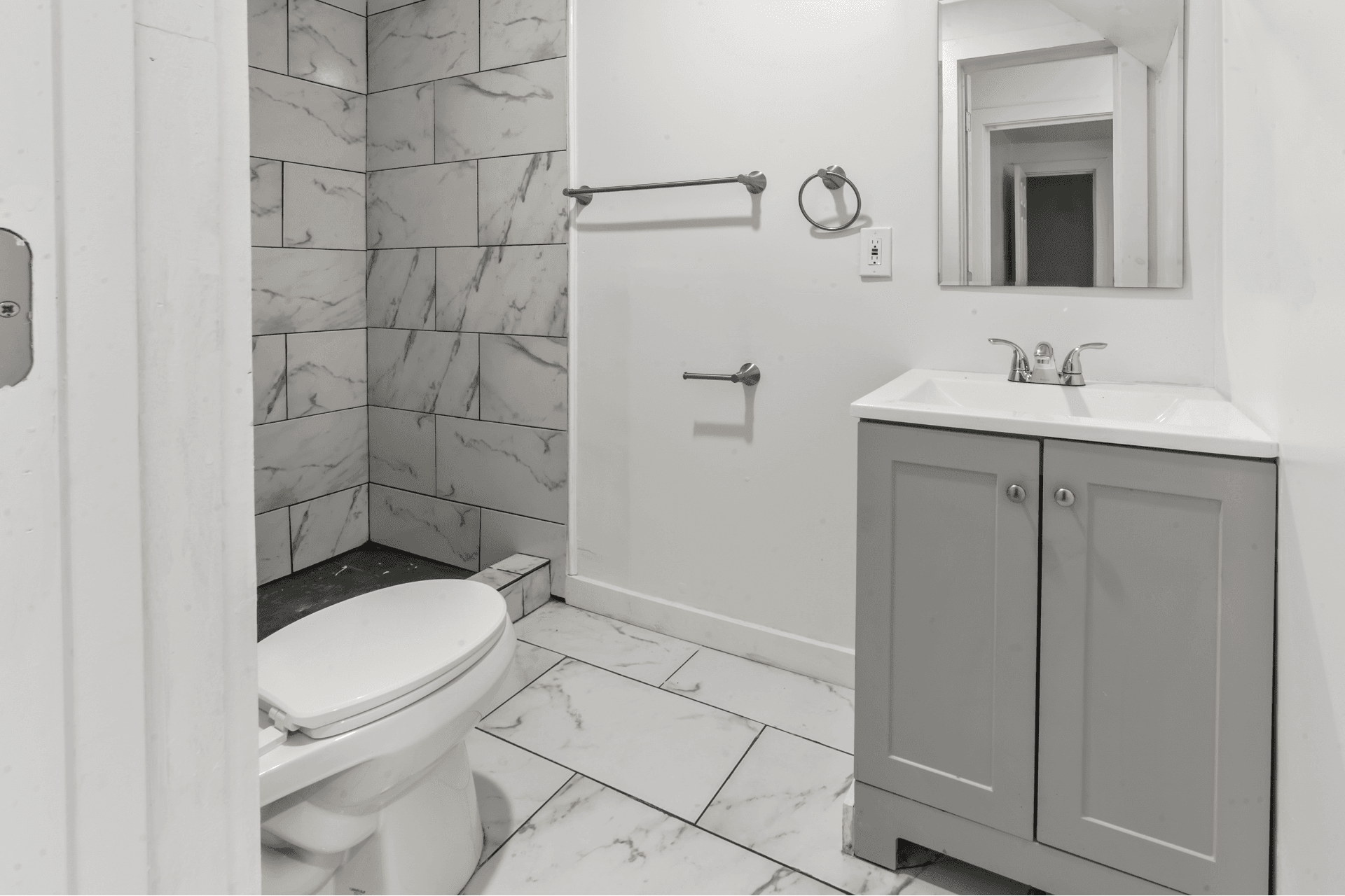 Bathroom with marble-tiled shower on the left, white toilet in foreground, and gray vanity with sink on the right.