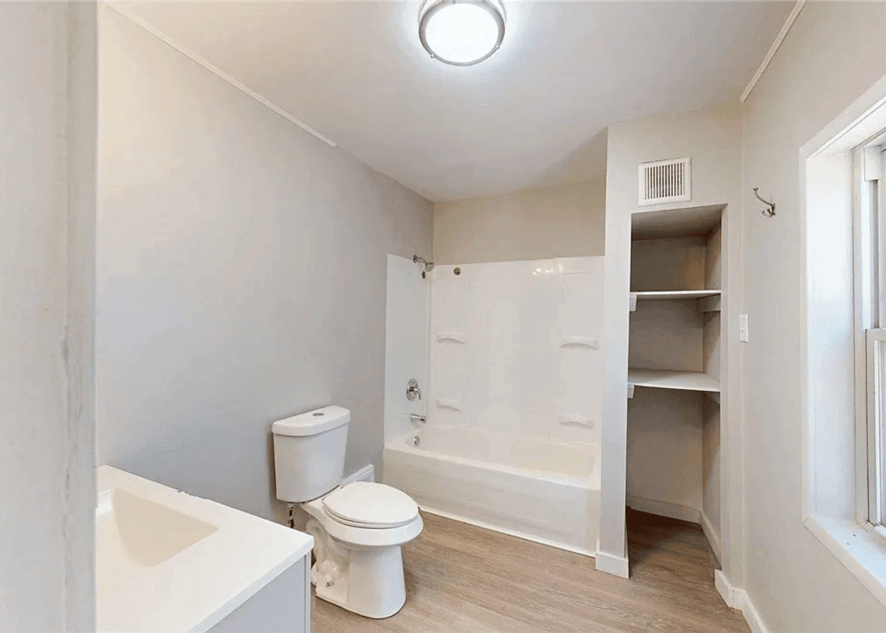 Bright bathroom with a white tub/shower combo, toilet, and open shelving unit beside a window on the right.