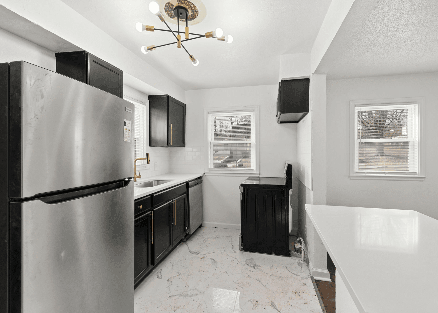 Modern kitchen with black cabinets, stainless steel refrigerator, and white marble-like floor. A gold faucet and stove area visible by windows.